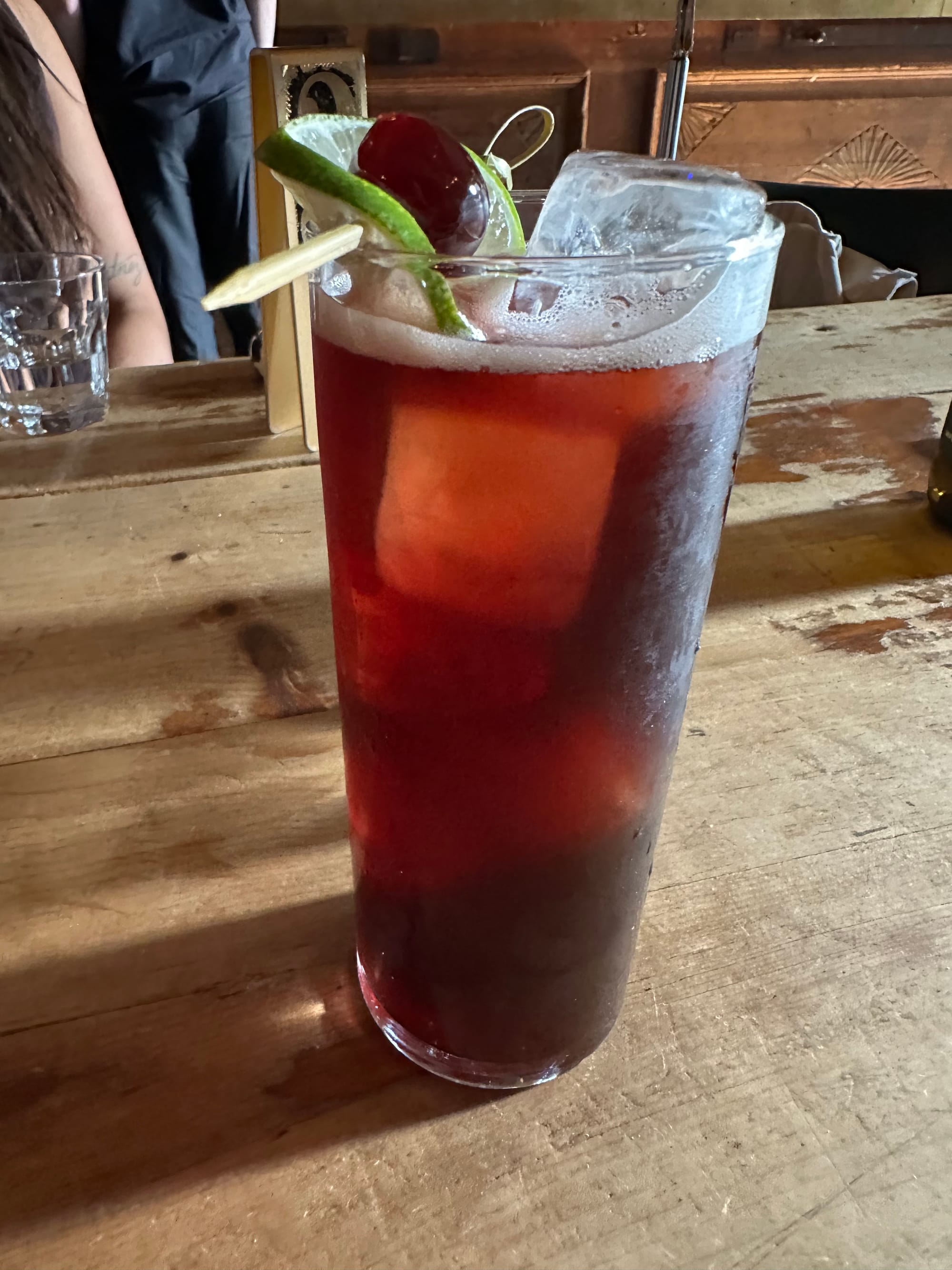 Highball glass on a wooden table. The drink is a reddish cola. The very cubic ice cubes are visible. It's garnished with a thin slice of lime wrapped around a cherry.