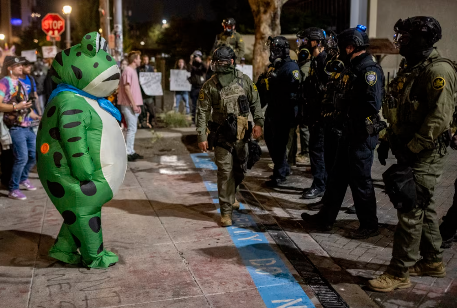 A person in an inflatable frog costume, heroically standing face to face with several federal agents.