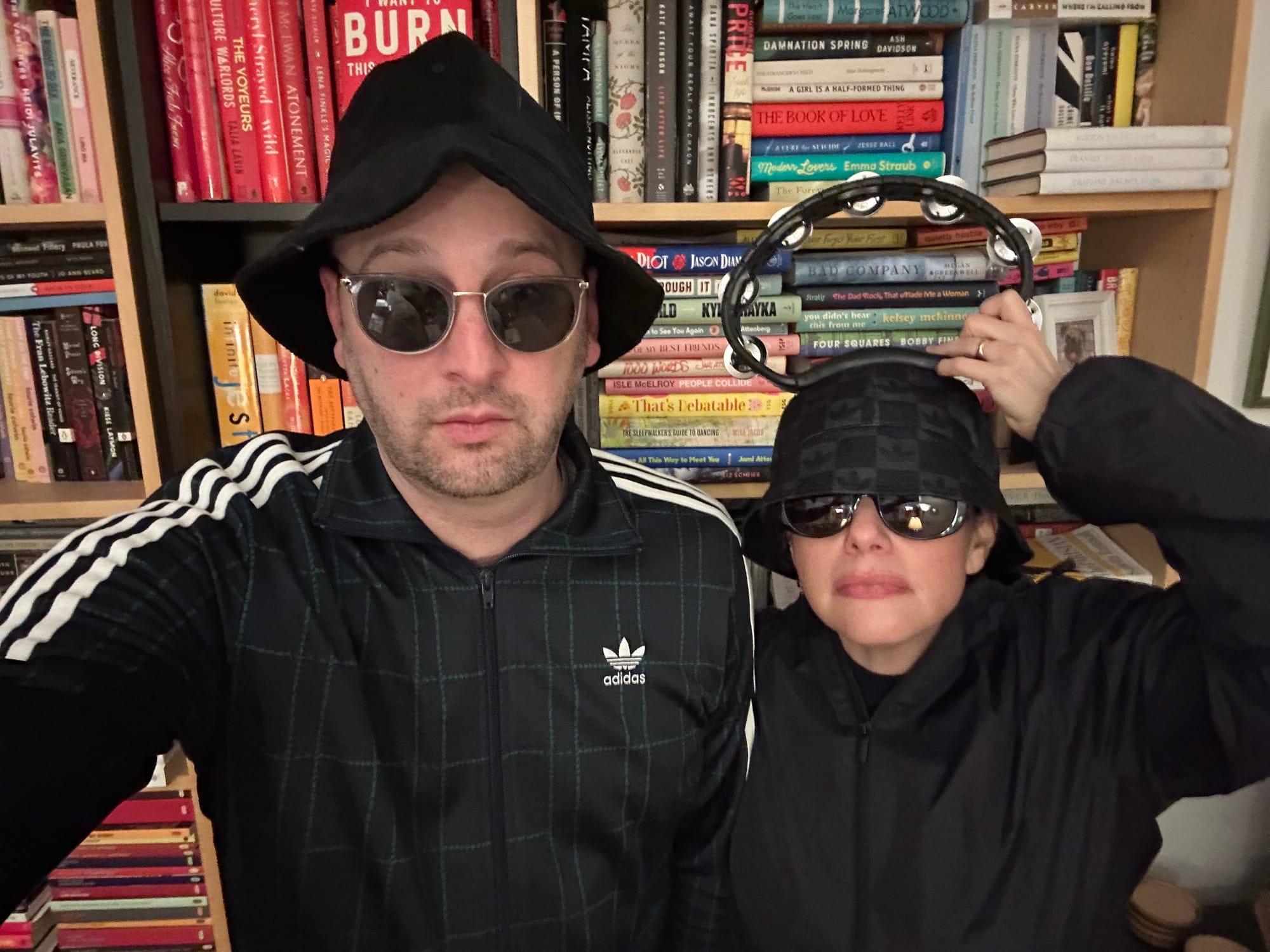 Me and Maris dressed as Oasis in bucket hats in front of Maris's bookshelf. She has a tambourine on her head, Liam Gallagher style.