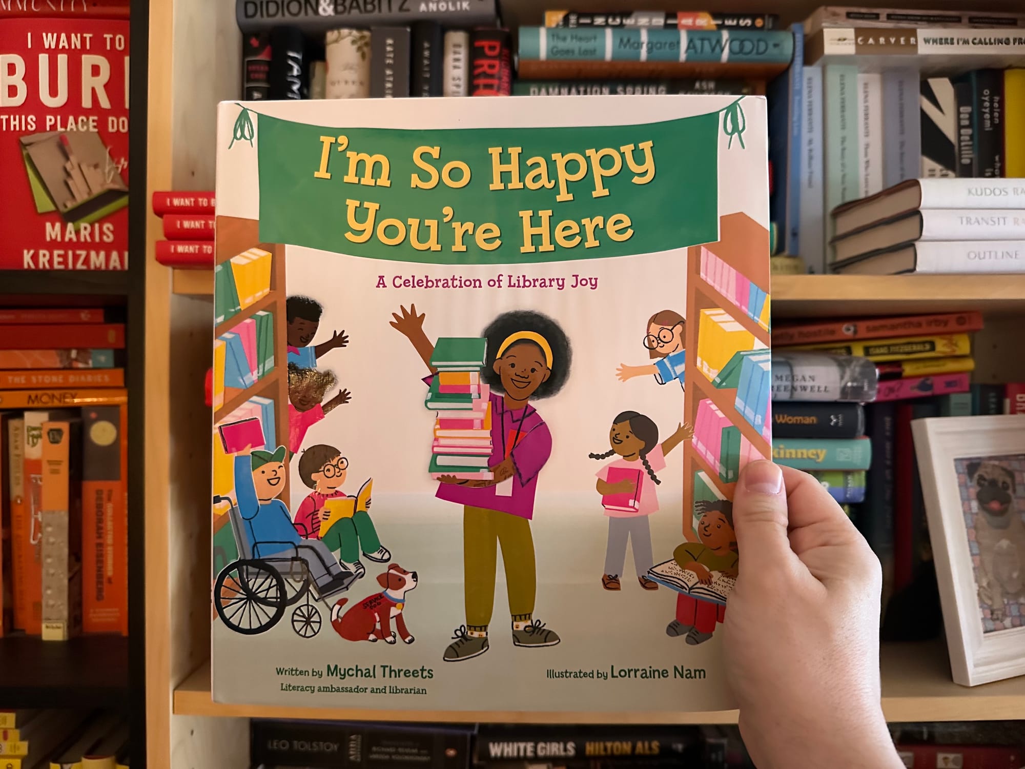 I'm So Happy You're Here: A Celebration of Library Joy by Mychal Threets in front of our bookshelf.
